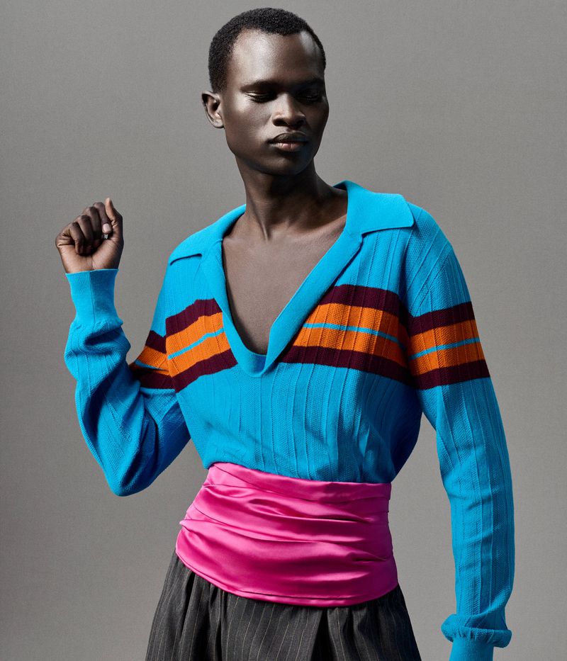 Studio shot of a black male model in a blue, orange, and maroon striped long-sleeve shirt, pink sash, and gray pinstripe pants on grey background.