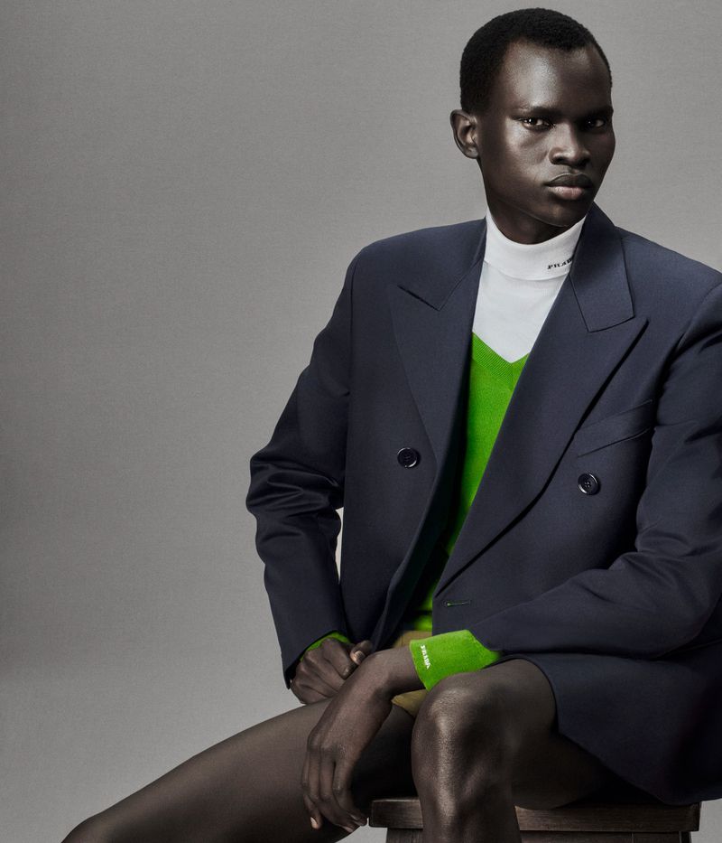 Model wears a dark blue suit jacket with a bright green sweater, posing while sitting on a stool in a studio with gray background.