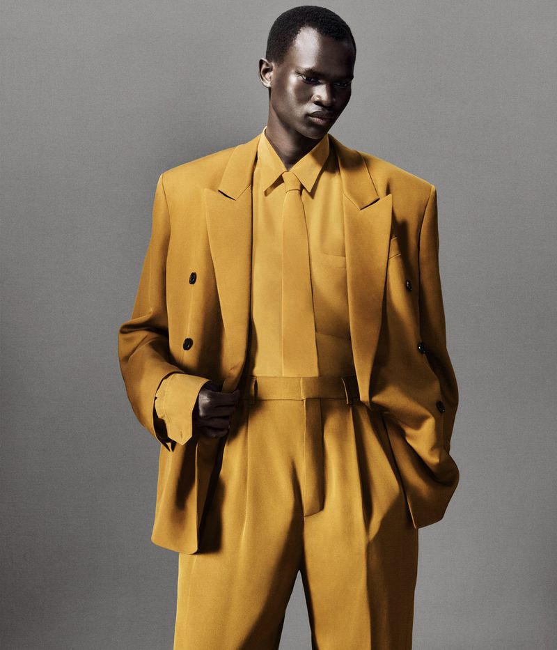 Adult male in a mustard-yellow double-breasted suit with a tie posing in a studio with a grey backdrop looking towards the camera.