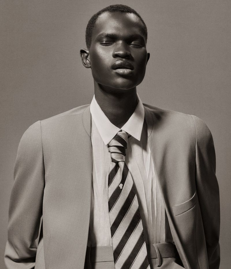 Portrait of a Black male model with closed eyes wearing a gray suit and tie against a solid gray backdrop in a studio setting.