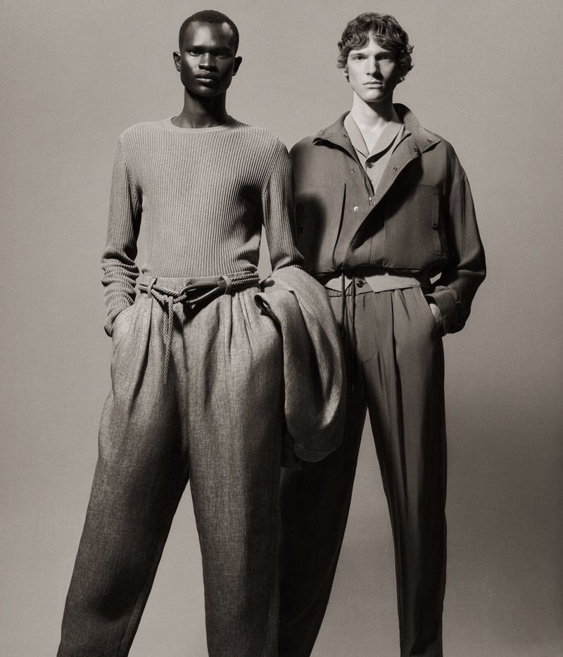 Two adult male fashion models pose in neutral-toned knit sweaters and trousers against a grey backdrop in a studio shot.