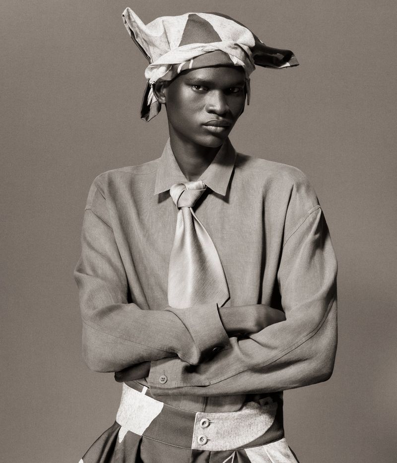 Portrait of a young black man in a light-colored shirt, tie, and head wrap, posing with crossed arms against a grey background.