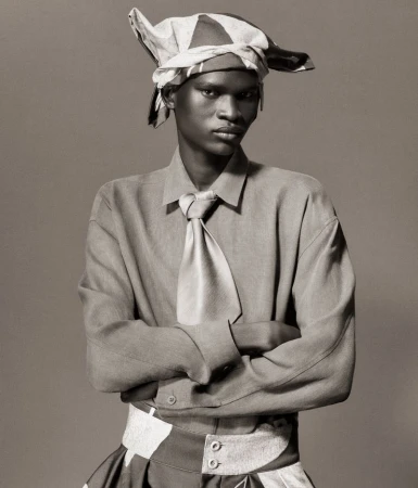 Portrait of a young black man in a light-colored shirt, tie, and head wrap, posing with crossed arms against a grey background.