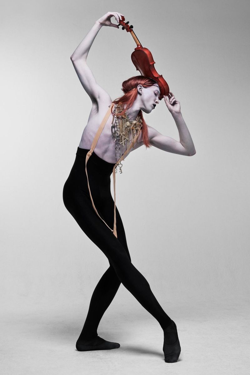 Artistic full shot of pale adult holding a violin overhead, wearing black tights and gold jewelry, against a grey studio background.