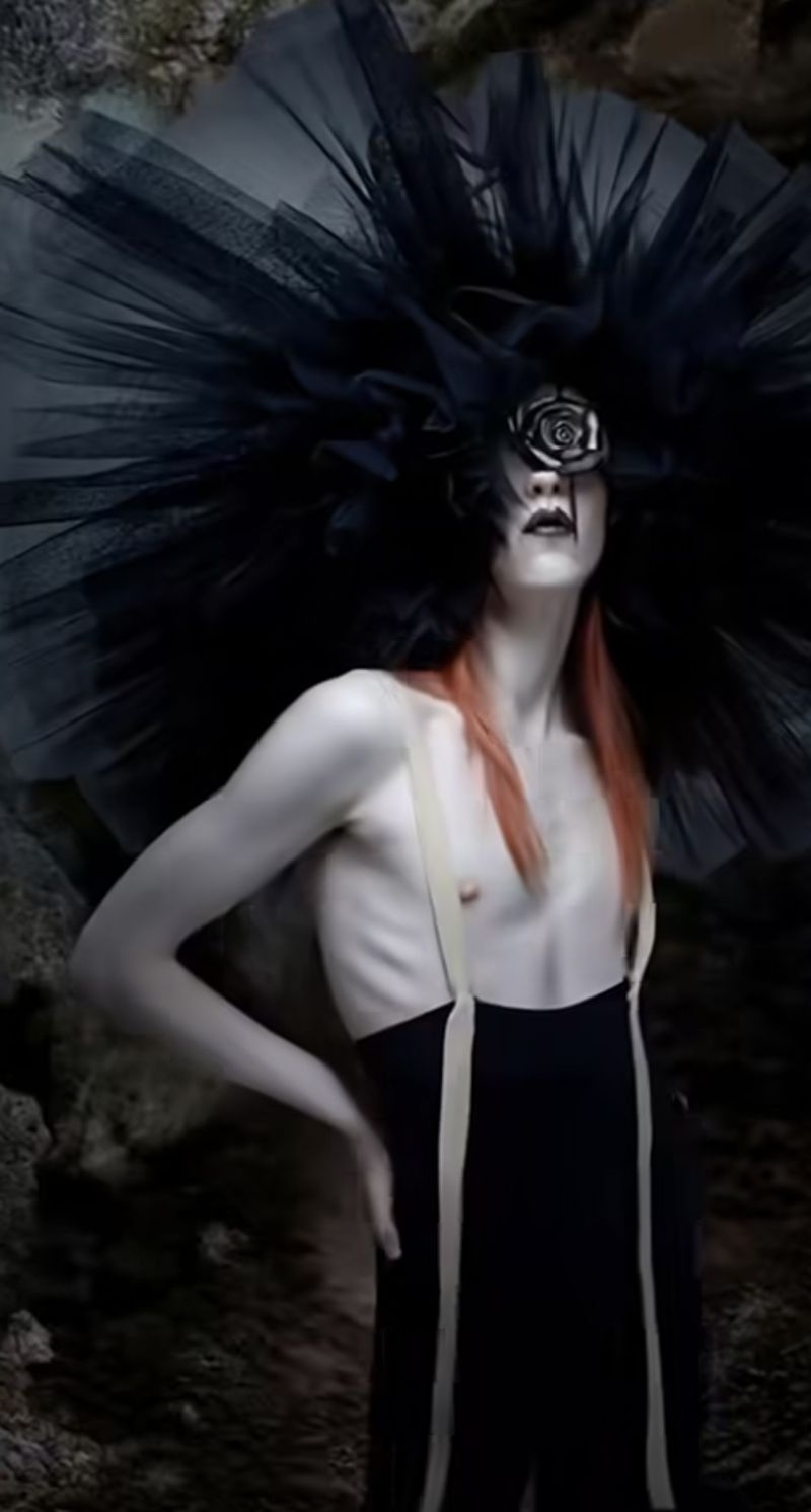 Person wears black formal wear, avant-garde black tulle hat with floral accent in studio, stone-like backdrop, red hair, fair skin. 