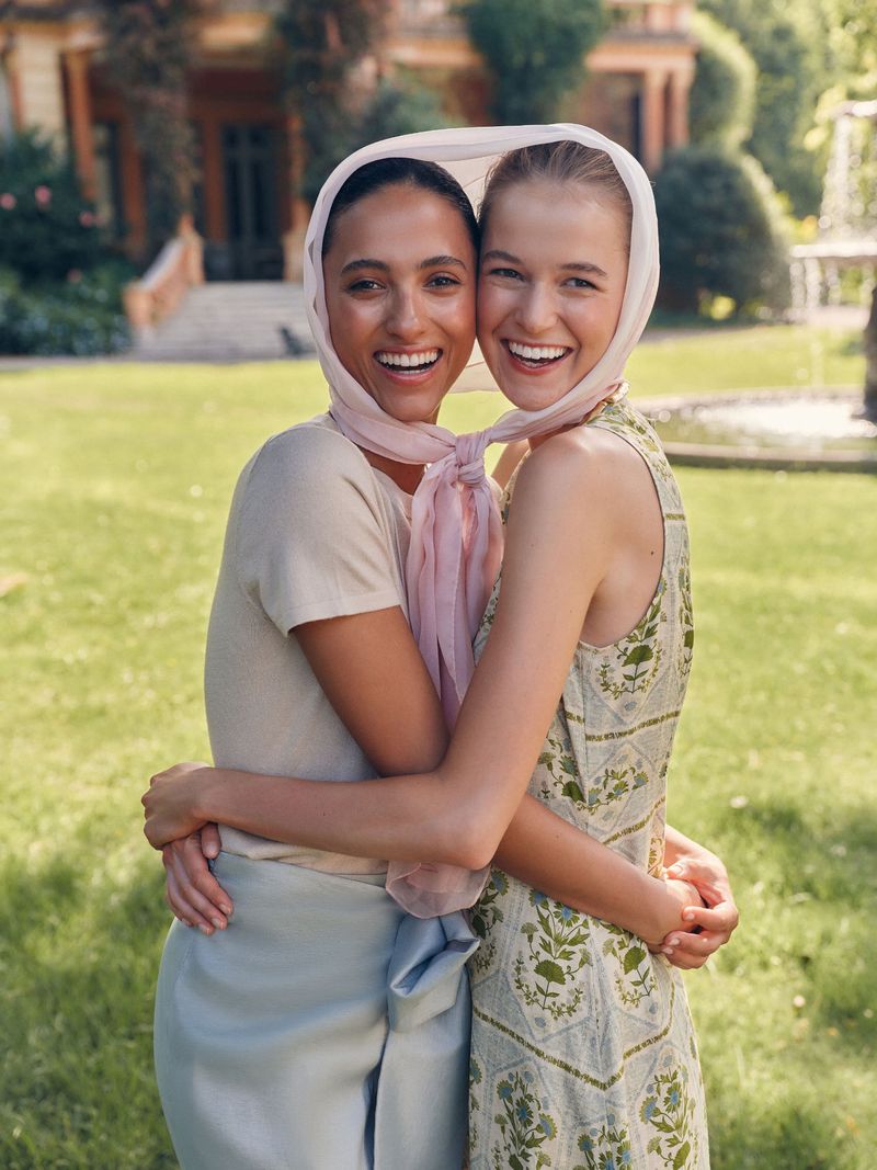 Two smiling women embrace in a sunny outdoor setting, wearing headscarves and light summer dresses, with a building and lawn background.