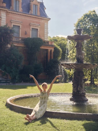 Young woman in a dress sits at the edge of a fountain in a garden near a building with hands up, enjoying a sunny day.