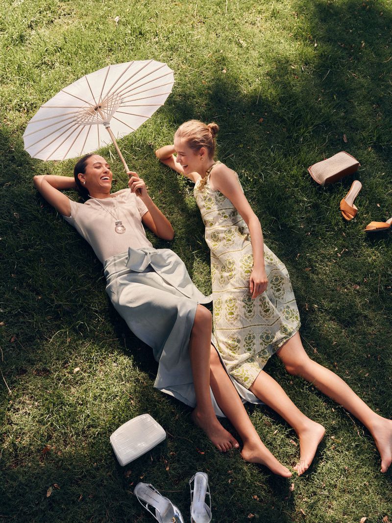 Two young women lie on the grass in the sun with shoes and purses scattered around them. One holds a white parasol.