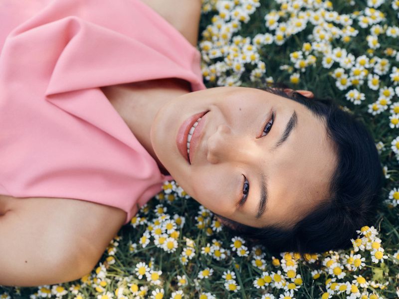 Young woman with dark hair lies in a field of daisies, smiling in a pink top, relaxed and content in a close-up shot on a sunny day.