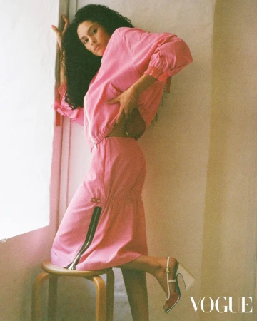 Fashion shot of woman in a pink two-piece outfit posing next to a stool in front of a window with the VOGUE logo in the corner.