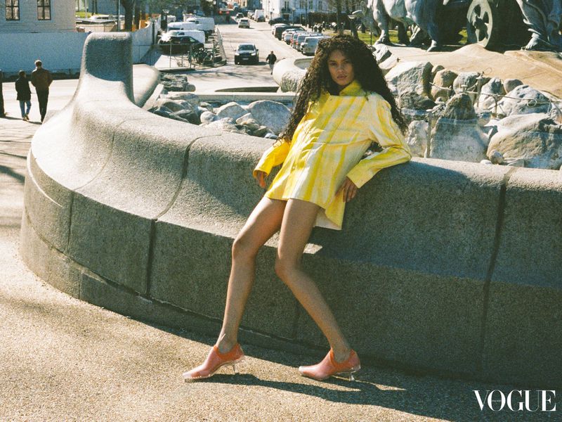 Brunette woman posing on a concrete ledge wearing yellow patterned jacket and orange shoes outdoors on a city street on a sunny day.