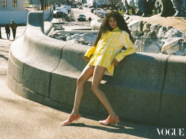 Brunette woman posing on a concrete ledge wearing yellow patterned jacket and orange shoes outdoors on a city street on a sunny day.