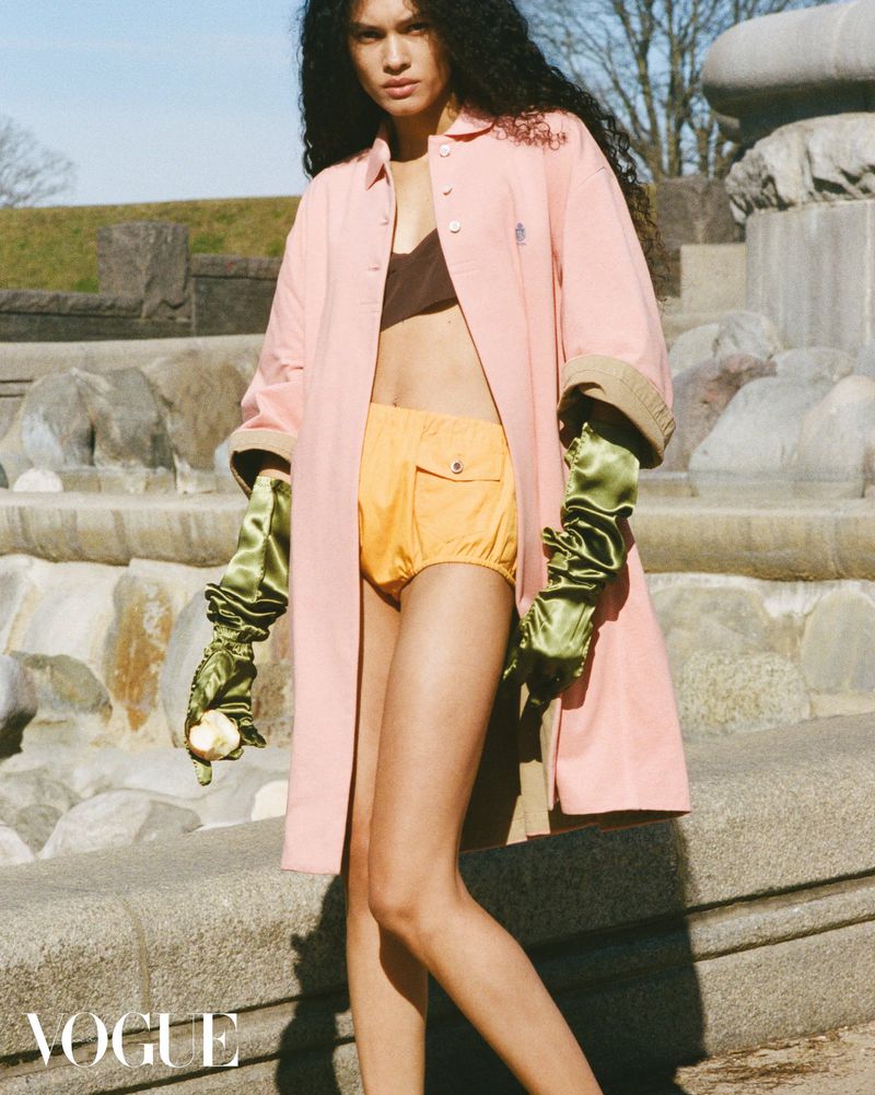 Fashion shot of a young adult woman in a pink overcoat, yellow shorts, and green gloves posing in front of a stone fountain.