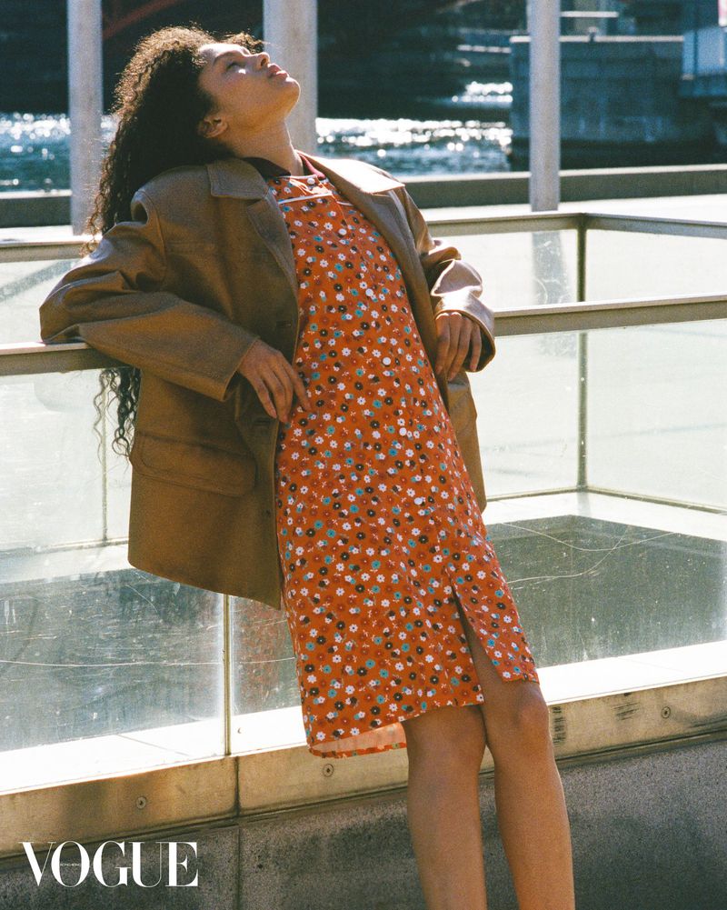 Woman with curly hair in a brown coat and red floral dress leans on a railing with a body of water and buildings in the background.