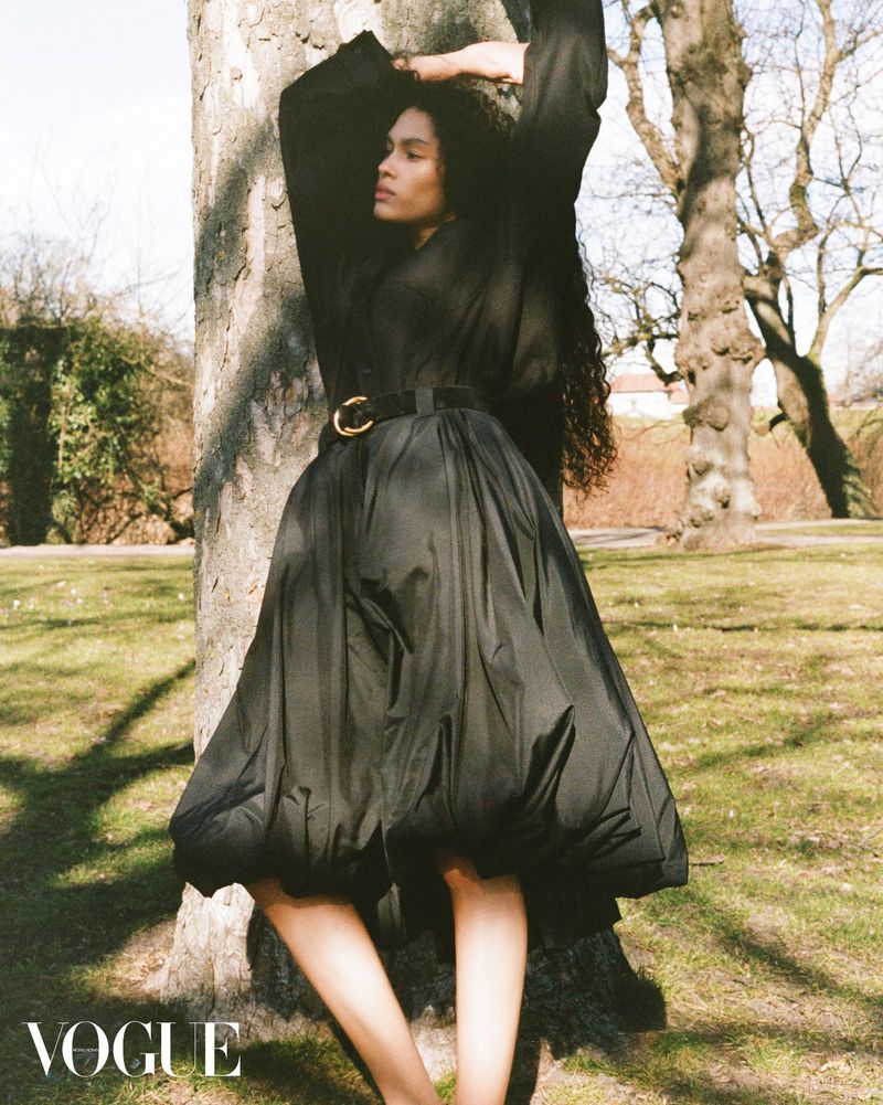 An adult woman with curly hair leans against a tree in a park. She is wearing a black dress with a belt, outdoors in natural sunlight.