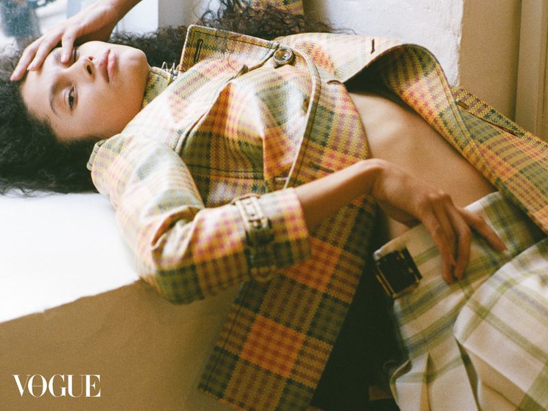 Fashion editorial shot of a woman in a plaid jacket and skirt, reclining against a bright window with natural light and Vogue logo.