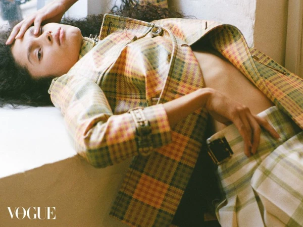 Fashion editorial shot of a woman in a plaid jacket and skirt, reclining against a bright window with natural light and Vogue logo.