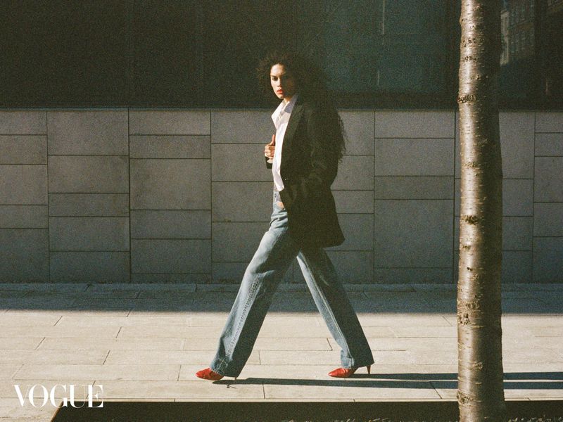 An adult female fashion model in a blazer, jeans, shirt, and heels walks on a city sidewalk next to a building and a tree trunk.