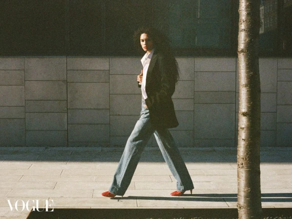 An adult female fashion model in a blazer, jeans, shirt, and heels walks on a city sidewalk next to a building and a tree trunk.