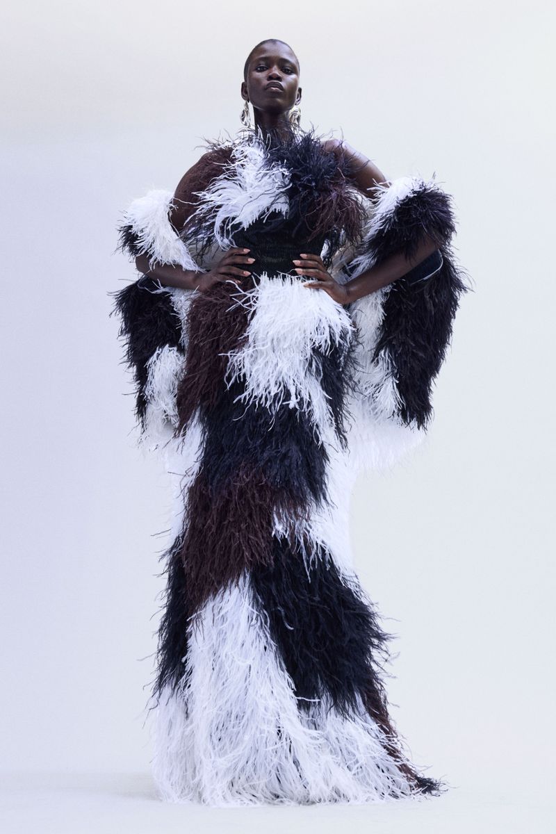 Elegant, full-length studio shot of a Black female model in a feathered black, white, and brown gown with statement earrings, hands on hips.