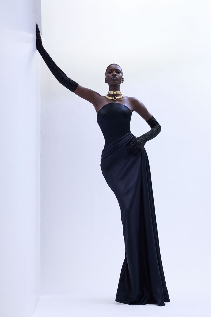 Model in a black strapless gown with long opera gloves posing against a white studio wall for Naeem Khan
