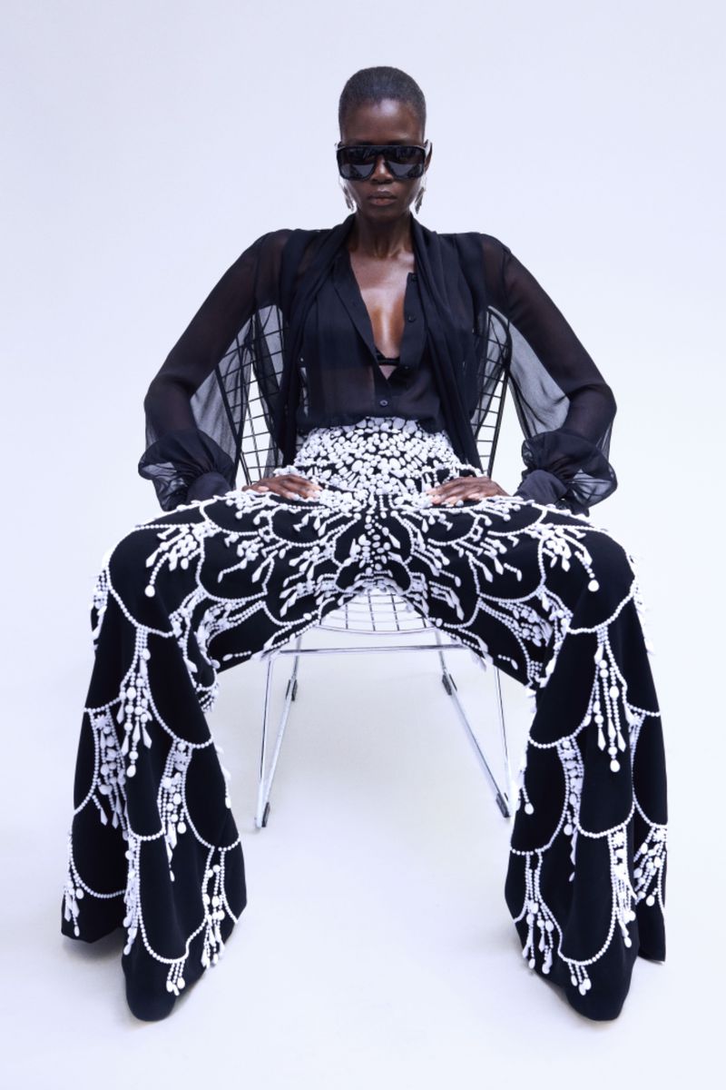 A dark-skinned woman in a black top and patterned pants, seated in a chrome chair against a white backdrop, modelling fashion.