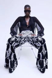 Model Ajah Angau Jok in black sheer blouse and heavily embellished trousers seated on a wire chair against white seamless background