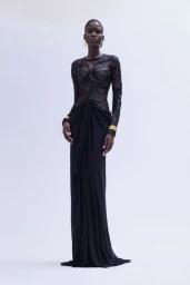 Model wearing a sheer black beaded long-sleeve gown with gold jewelry against a white backdrop