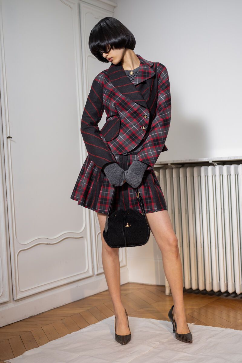 Woman in plaid blazer and skirt holding a purse, wearing heels and gloves, posing indoors against a white wall and radiator.