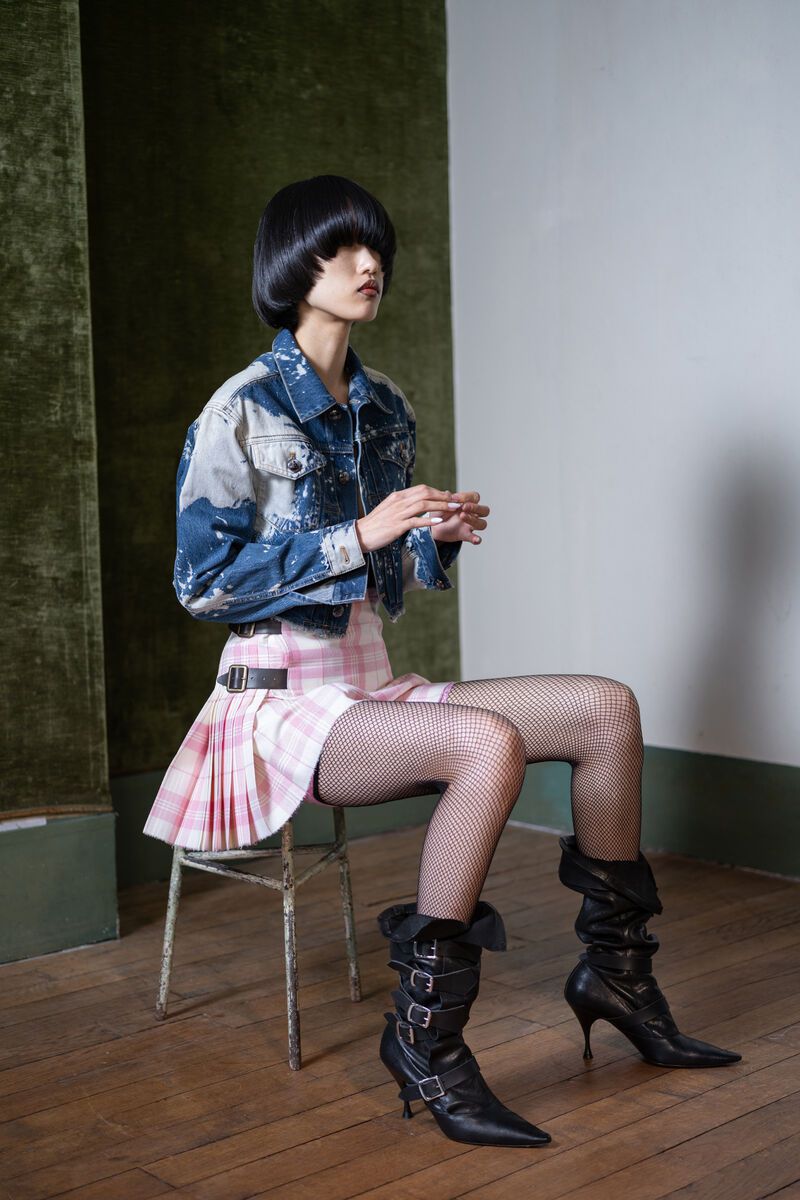 Young woman with a blunt bob haircut sits on a metal chair in a blue denim jacket and pink plaid skirt against a neutral backdrop.
