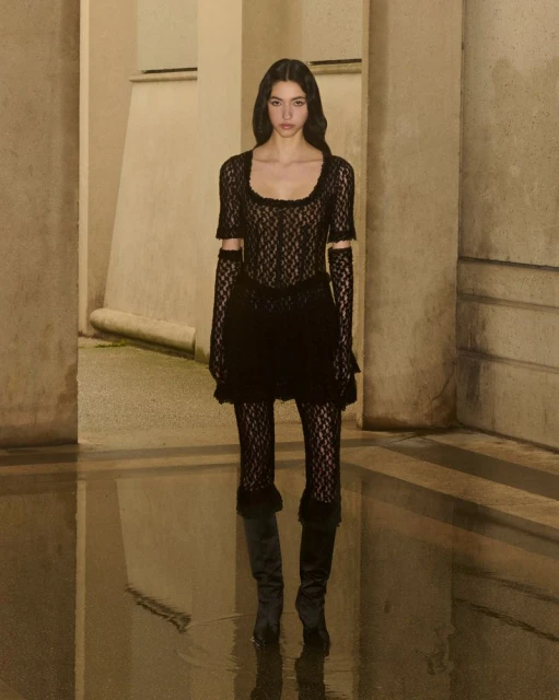 A woman in a black lace dress and boots stands indoors with a reflective floor and light beige columns in the background.