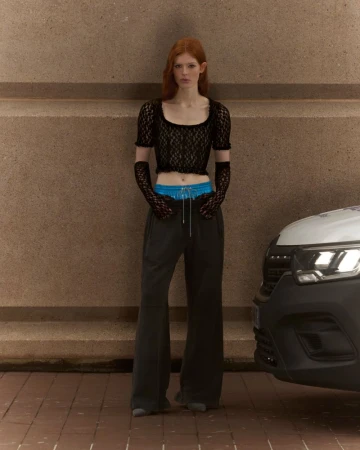 Fashionable woman in lace top and sweatpants poses against a neutral wall and delivery vehicle in an urban setting; full body shot.