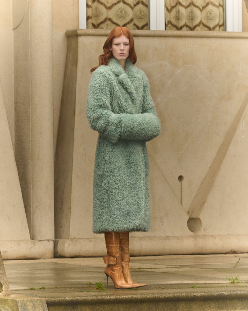 A ginger-haired woman stands tall, wrapped in a cozy green, faux fur coat and tan boots, posing against a neutral-toned building exterior.