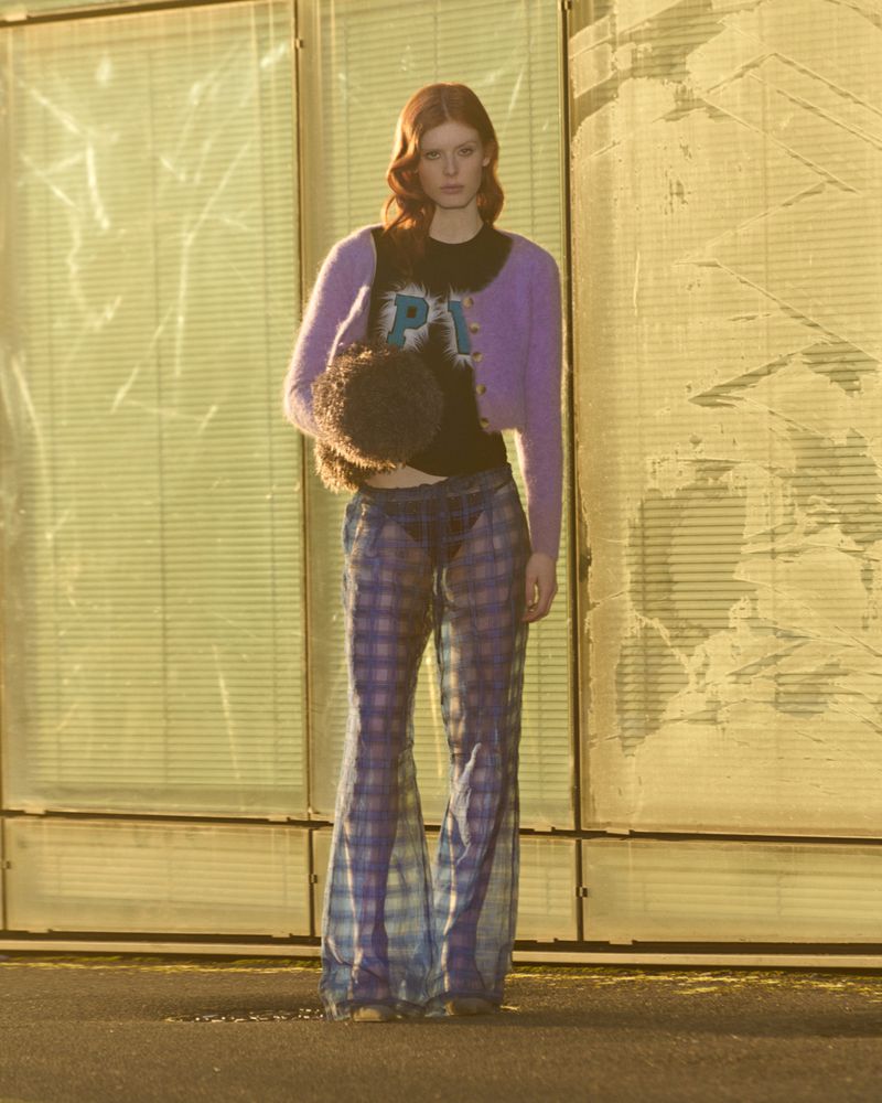 Redhead model poses in front of corrugated background wearing a black logo T-shirt, purple cardigan, plaid pants, and carrying a fur purse.