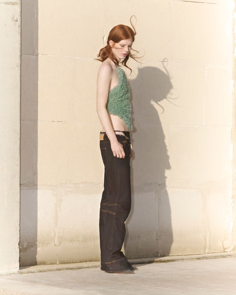 A slender redhead woman is posing in a studio, wearing a halter top and dark jeans with an open area around the hips, bathed in soft light.