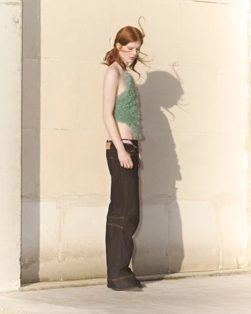 A slender redhead woman is posing in a studio, wearing a halter top and dark jeans with an open area around the hips, bathed in soft light.