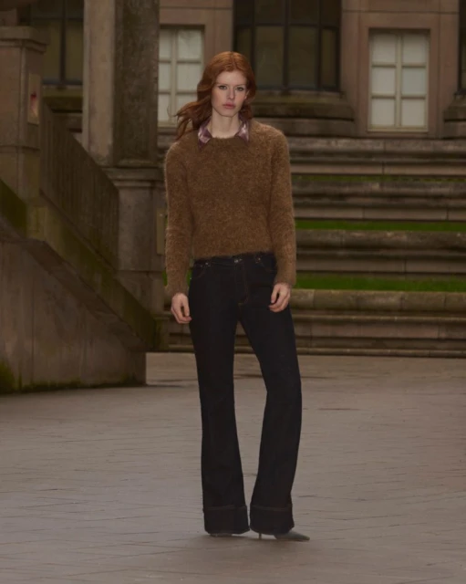 A redhead woman in brown sweater and black jeans poses in front of a building with stone columns and stairs in an outdoor setting.
