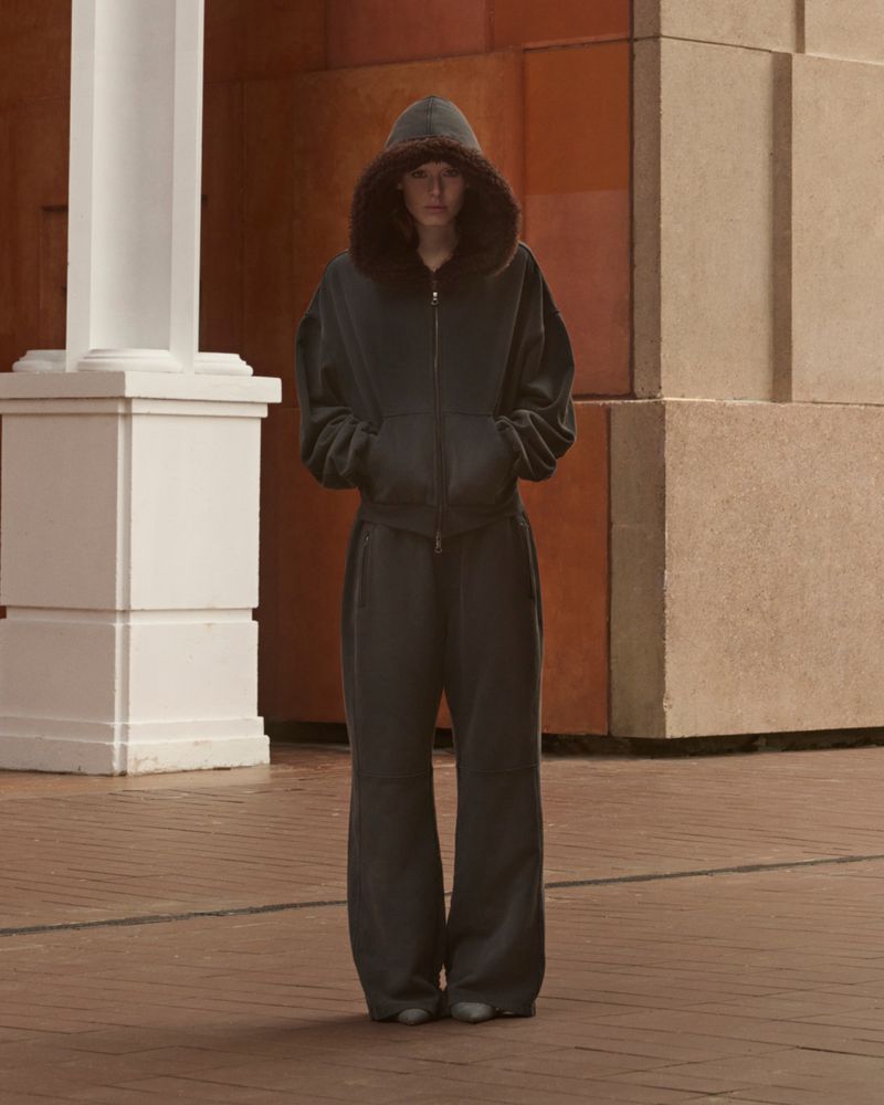 Woman wearing a dark grey hooded tracksuit standing in an open space with geometric wall and floor tiles and white pillars.