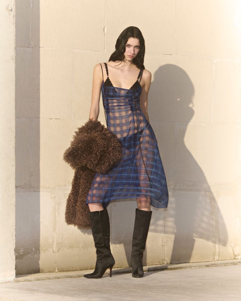 Fashion model in a semi-sheer blue plaid dress and boots, holding a fur jacket, posing in front of a neutral wall, full-length shot.