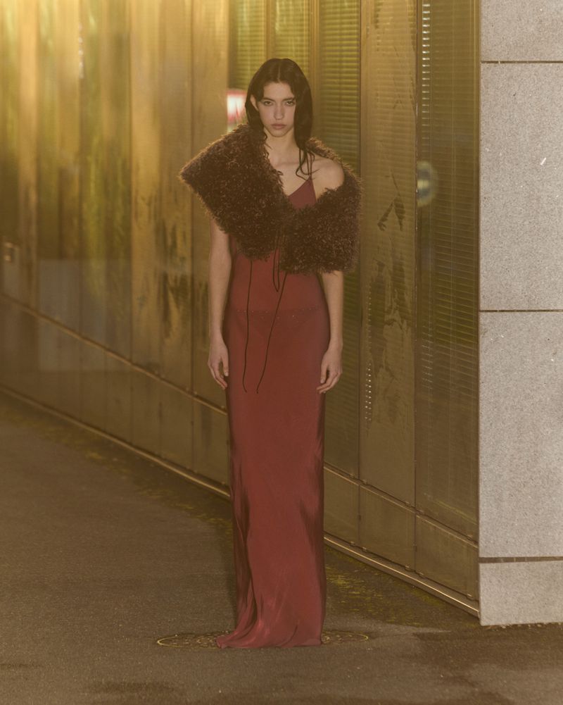 An adult woman with long brown hair poses in a long, tight, sleeveless maroon dress with a brown fur shawl in an indoor hallway.