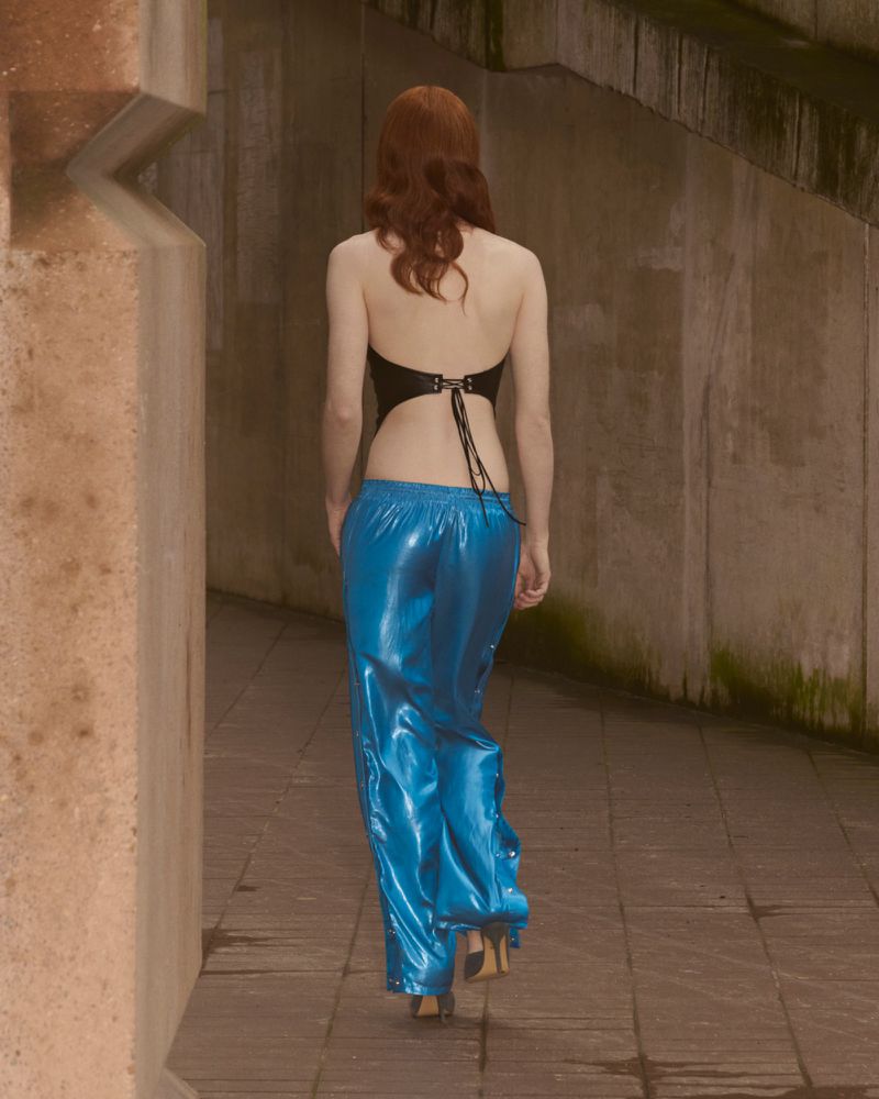 Woman walking away wearing a halter top and shiny blue pants on a stone pathway between concrete walls. Fashion or editorial shot.