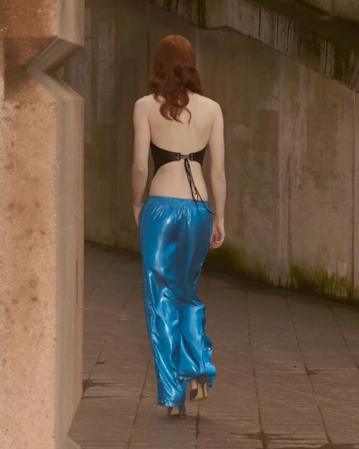 Woman walking away wearing a halter top and shiny blue pants on a stone pathway between concrete walls. Fashion or editorial shot.
