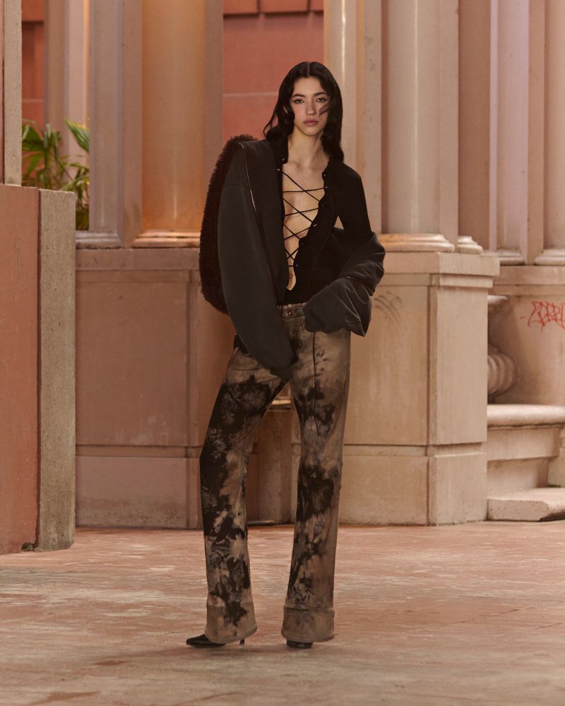 Fashionable woman in a lace-up black top and tie-dye pants posing in a neutral-toned outdoor setting, showcasing stylish apparel.