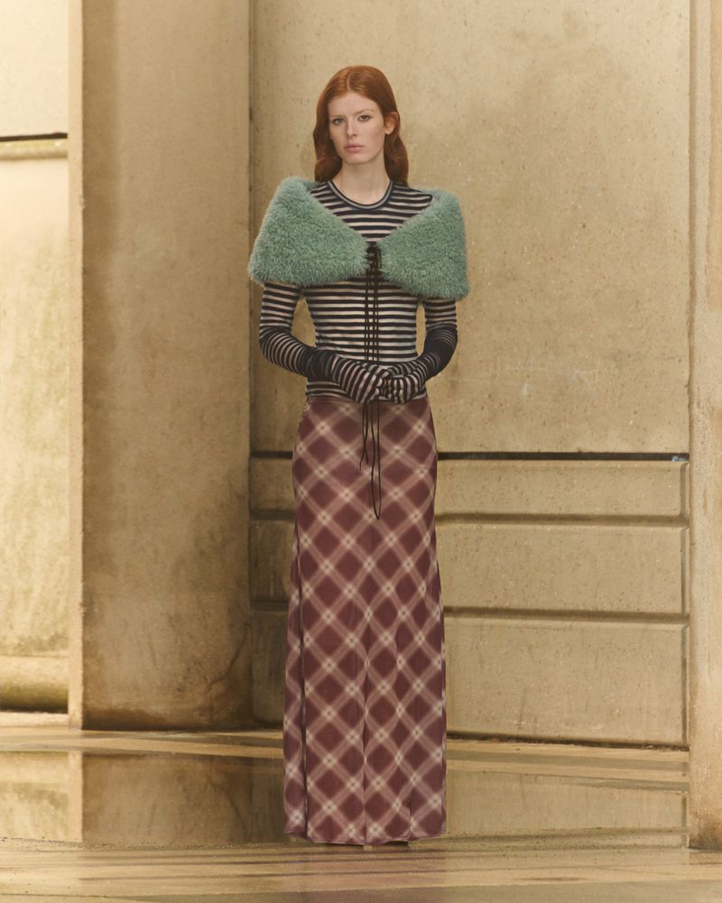 A red-haired woman in a long plaid skirt, striped top and fluffy green shawl poses in front of a tan wall. Fashion shot, studio lighting.