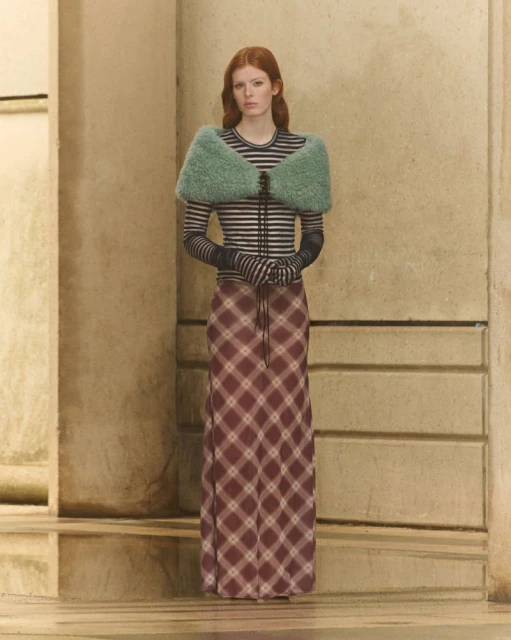 A red-haired woman in a long plaid skirt, striped top and fluffy green shawl poses in front of a tan wall. Fashion shot, studio lighting.