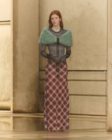 A red-haired woman in a long plaid skirt, striped top and fluffy green shawl poses in front of a tan wall. Fashion shot, studio lighting.