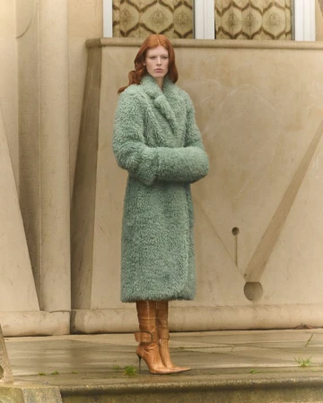 Model with red hair wearing a sage green fluffy coat and brown boots outdoors