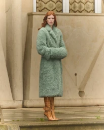 Model with red hair wearing a sage green fluffy coat and brown boots outdoors