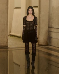 Model wearing a black lace dress and knee-high boots standing on a wet reflective pavement between stone architectural pillars.