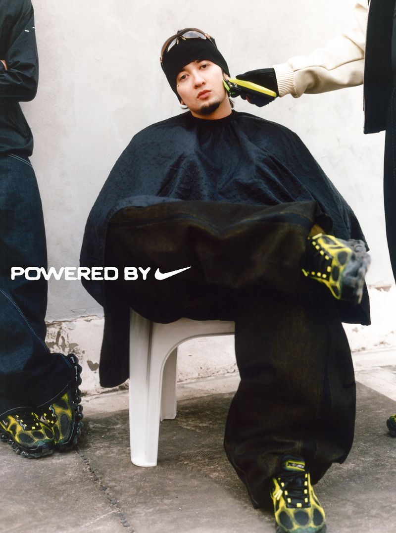 Man in a black cape getting a haircut outdoors with black and yellow sneakers, jeans and black top, ‘Powered by Nike’ logo visible.